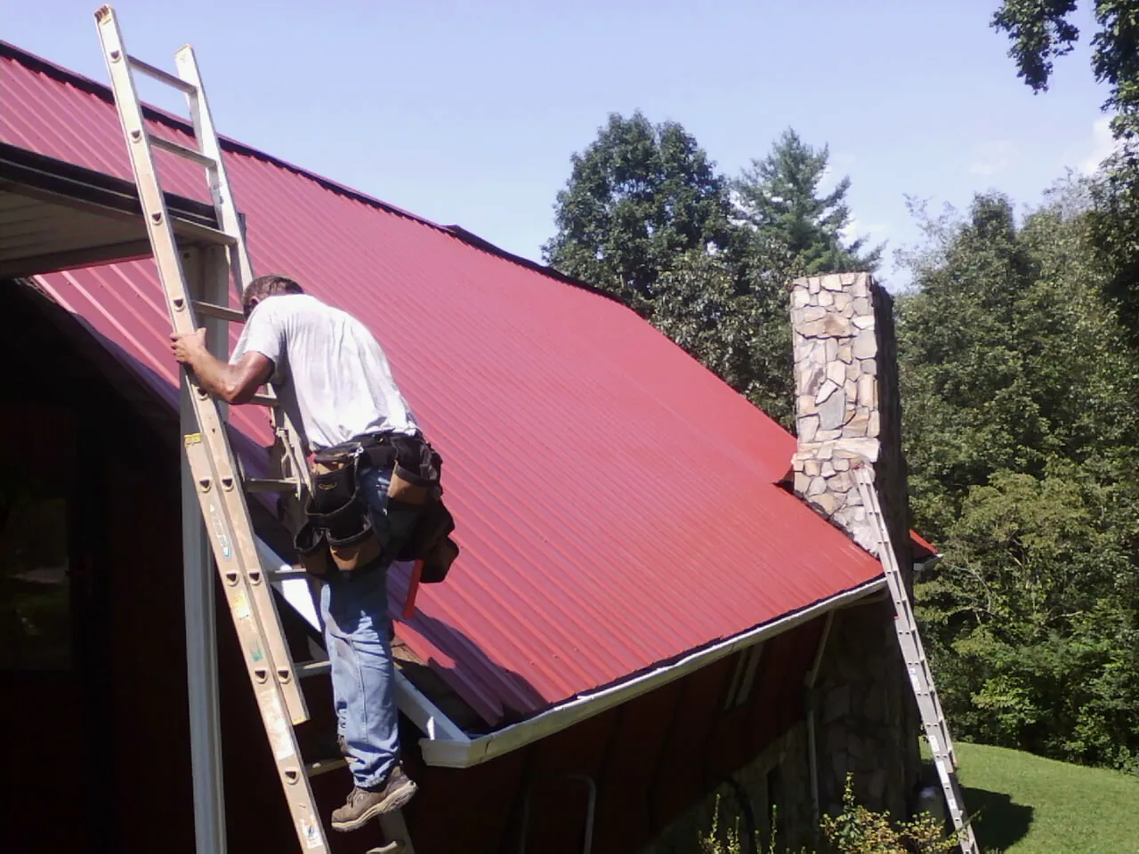 Roofer on a ladder beside a red metal roof and stone chimney — Soffit & Fascia Repair by Roofing Brothers in Cynthiana